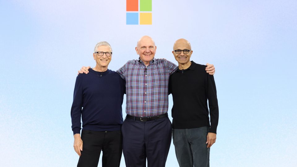 Three men with their arms around each other’s shoulders stand on stage in front of the colorful square Microsoft logo.